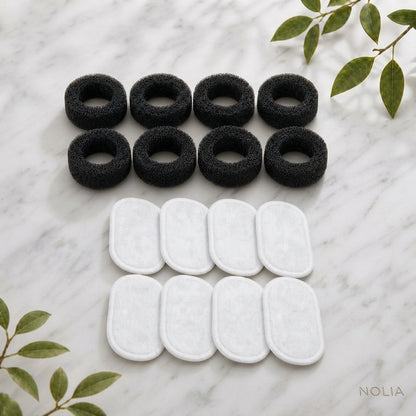 Lot complet de recharges pour fontaine à eau Nolia, présenté sur un fond en marbre clair avec des touches de feuillage, comprenant huit filtres en mousse noire de pré-filtration et huit filtres blancs haute performance.